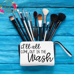 May include: White makeup bag with black trim and a zipper. The bag has the text "it'll all come out in the wash" printed on it. The bag is open and filled with a variety of makeup brushes.
