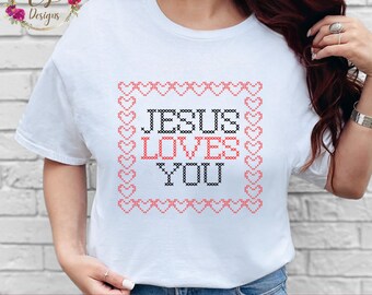 May include: White t-shirt with a cross-stitch design. The design features the words "JESUS LOVES YOU" in black and red, framed by a border of red hearts. The shirt is a casual style.