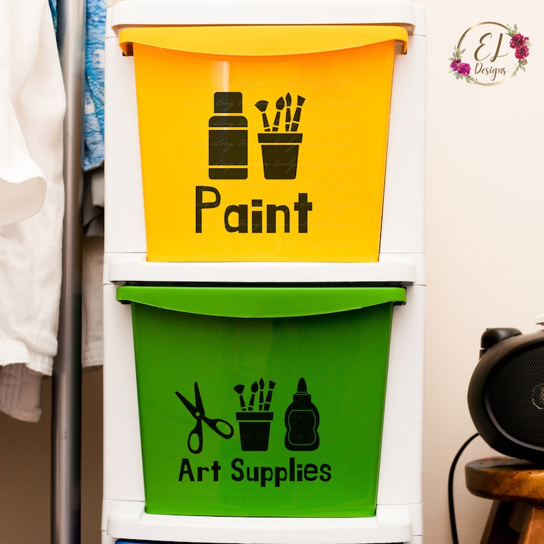 May include: Two yellow and green plastic storage bins with labels. The top bin is yellow and says "Paint" with a paint bottle and paintbrushes in a cup. The bottom bin is green and says "Art Supplies" with scissors, paintbrushes in a cup, and a bottle of glue.