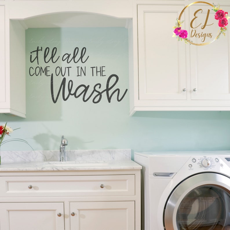 May include: A white laundry room with a white cabinet and a white washing machine. The wall is light blue and has a black vinyl decal that says "it'll all come out in the wash".