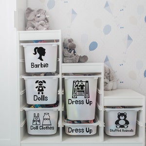 May include: White storage bins with black labels on a white shelf. The labels read "Barbie", "Dolls", "Doll Clothes", "Dress Up", "Dress Up", and "Stuffed Animals".