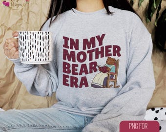May include: A gray sweatshirt with the text "In My Mother Bear Era" printed in maroon. A cartoon illustration of a brown bear sitting in a rocking chair and reading a book is also printed on the sweatshirt.