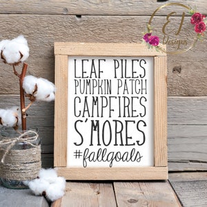 Op de afbeelding: Een houten bord met de woorden "LEAF PILES, PUMPKIN PATCH, CAMPFIRES, S'MORES, #fallgoals" in zwarte letters. Een katoenen tak in een glazen pot gewikkeld met touw staat links. De achtergrond is verweerd hout.