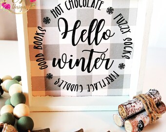 May include: White wooden sign with a black and white buffalo plaid background. The sign says "Hello winter" in a black script font. The words "hot chocolate", "good books", "fireplace", "cuddles", "fuzzy socks" are written in a circle around the main text.
