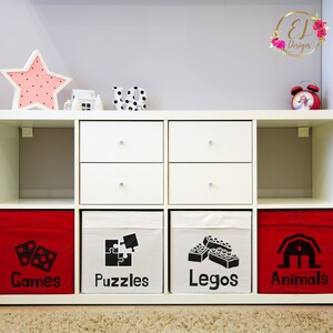 Puede incluir: Estanter&iacute;as blancas en forma de cubo con contenedores de almacenamiento rojos y blancos etiquetados como "Juegos", "Rompecabezas", "Lego" y "Animales". Cada contenedor tiene un icono negro que representa la categor&iacute;a.