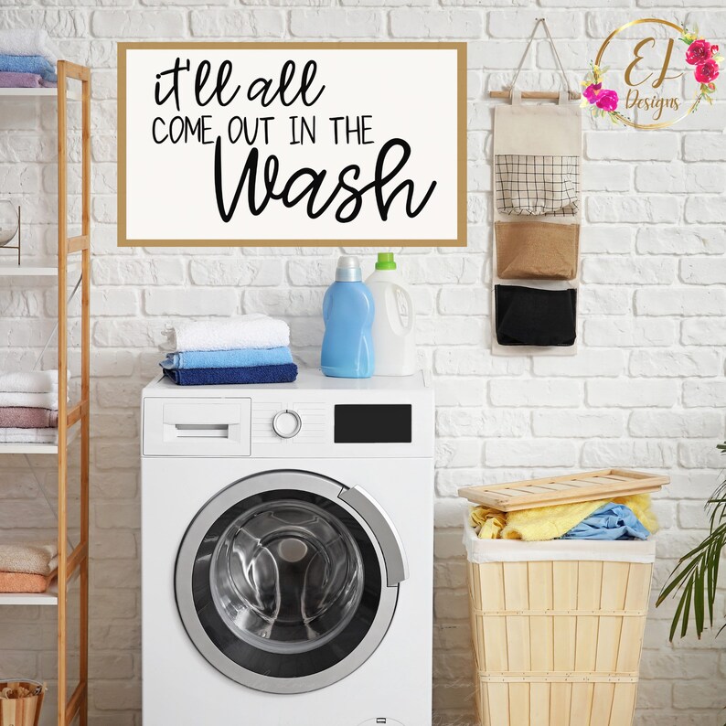 May include: White washing machine with a black digital display and a silver door. A wooden shelf with towels is on the left. A sign with the text "it'll all come out in the wash" is on the wall above the washing machine. A wooden basket with folded laundry is on the right.