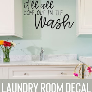 May include: A white laundry room with a white cabinet and a white sink. The wall is light blue and has a black vinyl decal that says "it'll all come out in the wash".