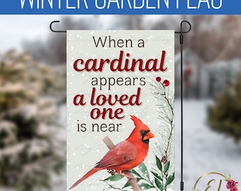 Op de afbeelding: Een wintertuin vlag met een rode kardinaal die op een tak zit. De vlag heeft een witte achtergrond met de tekst "Wanneer een kardinaal verschijnt, is een geliefde in de buurt".