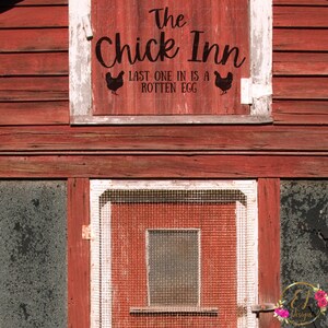 以下が含まれることがあります： 白い枠の窓のある赤い木製の納屋。窓には「The Chick Inn」と書かれた黒いビニール製の看板があり、ニワトリの黒いシルエットが2つと「Last one in is a rotten egg」という文字が書かれている。
