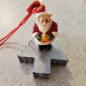 May include: A miniature Santa Claus figurine with a white beard, red suit, and a gold star, standing on a silver Christmas tree-shaped ornament. A red string is attached for hanging. Festive holiday decor.