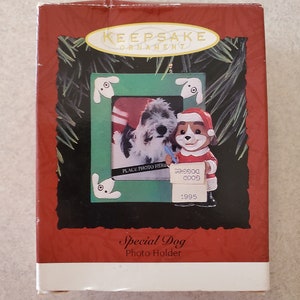 May include: A red and white keepsake ornament box featuring a photo frame with a dog wearing a Santa hat and a dog wearing a red scarf. The frame has a place to insert a photo and the text "Place Photo Here". The box also has the text "Special Dog Photo Holder" and "1995".