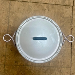 May include: A round, silver-colored cooking pot with a lid and two looped handles. The lid has a dark blue handle. The pot is made of metal and sits on a wooden surface. Kitchenware.