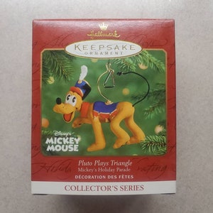 May include: A red box with a Hallmark Keepsake Ornament featuring Pluto playing a triangle. The ornament is part of Mickey's Holiday Parade and is a Collector's Series item.