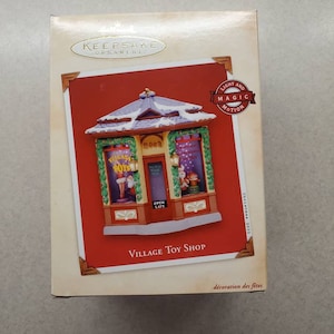 May include: Keepsake Christmas ornament box featuring a miniature Village Toy Shop. The red background highlights the shop's facade, with the year 2002 displayed. The box includes the text "Light and Magic Motion".