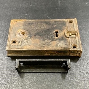 May include: An antique, rectangular metal lock with a keyhole and a separate black metal strike plate. The lock is heavily rusted, showing signs of age and wear. The lock has a square hole and two screw holes.
