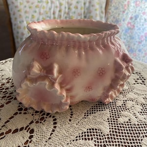 May include: A pink ceramic planter with a ruffled edge and bow details. The planter has a round shape and is decorated with small floral patterns. The overall design is vintage and delicate, suitable for small plants or flowers.
