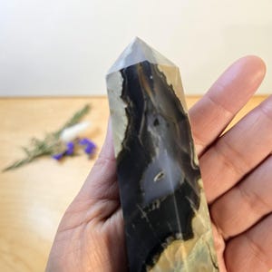 May include: A polished crystal obelisk with a pointed top. The stone features a mix of black, white, and tan colors with unique patterns. The obelisk is held in a hand, showcasing its size and shape. A decorative item for home decor.