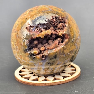 May include: A polished, spherical geode with a large cavity revealing a cluster of dark brown crystals. The exterior features a mix of brown, yellow, and gray hues. It rests on a decorative wooden coaster with a geometric design.