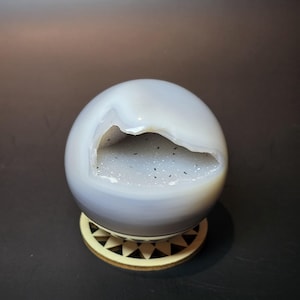 May include: A smooth, spherical light gray agate stone featuring a central geode cavity. The interior of the opening is lined with fine, sparkling white crystals speckled with tiny black inclusions, resting on a circular wooden stand.
