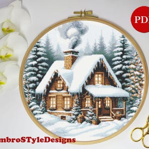 May include: A cross-stitch design of a snow-covered cabin in a winter forest, framed in a wooden embroidery hoop. The cabin has a snow-covered roof and chimney, with smoke rising. The text "EmbroStyleDesigns" is at the bottom.