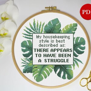 May include: Cross stitch pattern featuring a tropical leaf design with the text "My housekeeping style is best described as: THERE APPEARS TO HAVE BEEN A STRUGGLE".