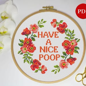 Könnte beinhalten: Ein weißer Stickrahmen mit einem Kreuzstich-Design aus roten Blumen und grünen Blättern, die den Text "HAVE A NICE POOP" umgeben.