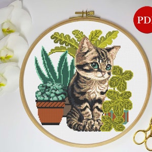 以下が含まれることがあります： 緑の植物の鉢に座っているかわいいトラ猫のクロスステッチパターン。子猫は緑色の目をしており、緑豊かな葉に囲まれています。