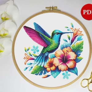 May include: A colourful cross stitch pattern featuring a hummingbird with green, blue and pink feathers, perched on a branch with pink and yellow flowers.