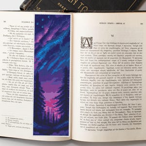 May include: A vibrant, pixelated bookmark featuring a colorful design of blues, purples, and pinks, resembling a stylized landscape or aurora borealis. The bookmark is placed in an open book with Greek text.