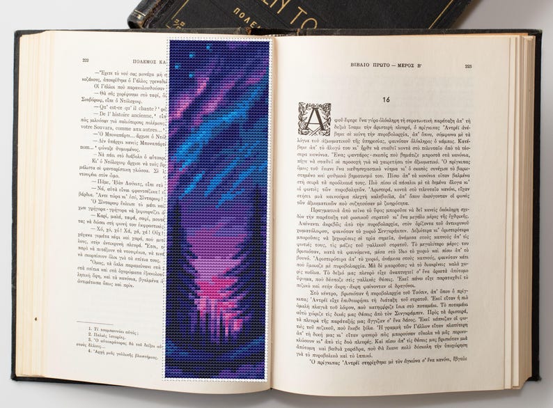 May include: A vibrant, pixelated bookmark featuring a colorful design of blues, purples, and pinks, resembling a stylized landscape or aurora borealis. The bookmark is placed in an open book with Greek text.