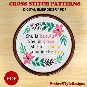May include: A cross-stitch pattern with the text "She is beauty, She is grace, She will punch you in the face" surrounded by a floral wreath. The design is in a round wooden frame, with the words "CROSS STITCH PATTERNS" at the top.