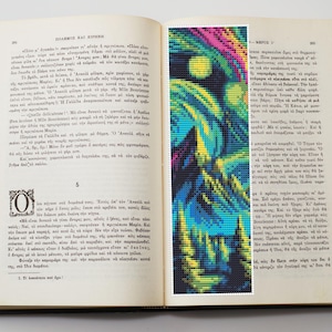 May include: A colorful, abstract bookmark featuring a pixelated design of a night sky with a full moon, and a forest. The bookmark is placed inside an open book with Greek text.