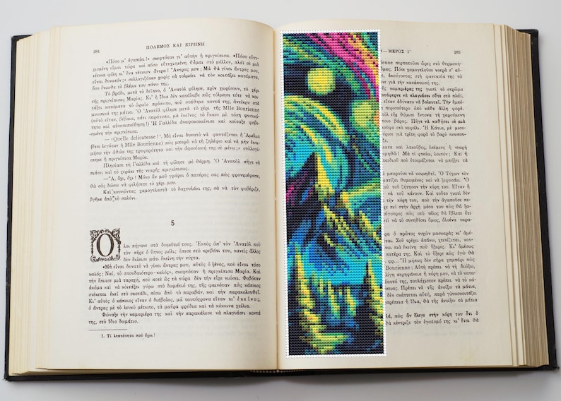 May include: A colorful, abstract bookmark featuring a pixelated design of a night sky with a full moon, and a forest. The bookmark is placed inside an open book with Greek text.
