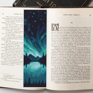 May include: A handmade bookmark featuring a cross-stitch design of the Northern Lights reflected in water. The bookmark is a rectangle with a dark blue background and teal and white accents. The bookmark is placed inside an open book.
