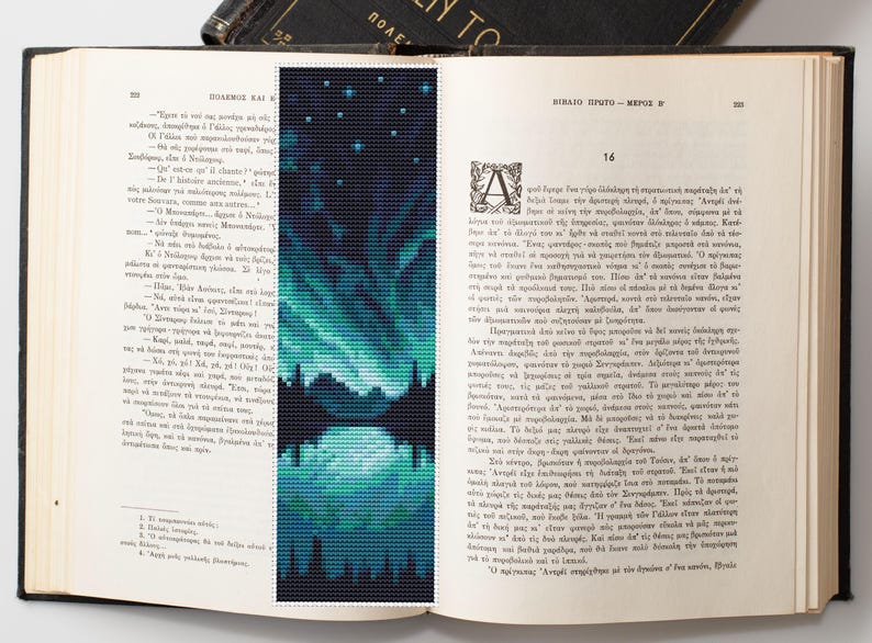 May include: A handmade bookmark featuring a cross-stitch design of the Northern Lights reflected in water. The bookmark is a rectangle with a dark blue background and teal and white accents. The bookmark is placed inside an open book.