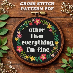 May include: A circular wooden embroidery hoop containing a black fabric cross stitch piece with the white text "other than everything, I'm fine" surrounded by a colorful floral wreath of pink, yellow, and green stitched flowers and leaves.