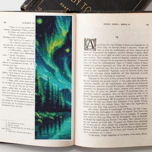 May include: A handmade bookmark featuring a pixelated design of an aurora borealis scene in shades of blue, green, and yellow. The bookmark is placed inside an open book with Greek text.
