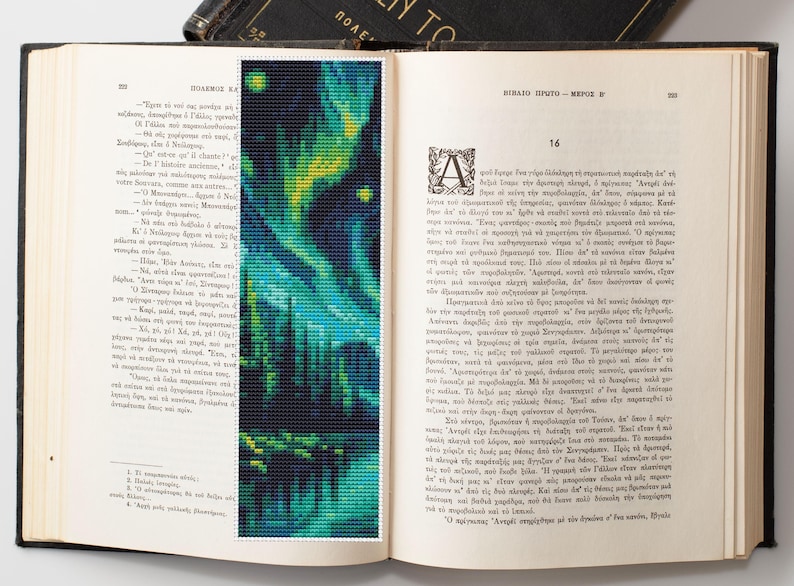 May include: A handmade bookmark featuring a pixelated design of an aurora borealis scene in shades of blue, green, and yellow. The bookmark is placed inside an open book with Greek text.