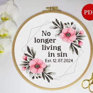 May include: A white embroidery hoop with a cross stitch design featuring pink flowers, black leaves, and the text "No longer living in sin Est. 12.07.2024".