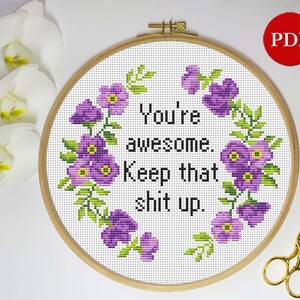 Puede incluir: Un bastidor de bordado de punto de cruz con el texto "You're awesome. Keep that shit up." rodeado de flores moradas y hojas verdes. Un par de tijeras doradas descansa en el lado derecho. Un icono PDF rojo está en la esquina superior derecha.