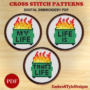 May include: Three cross stitch patterns featuring a green trash can with flames on top and the text "That's Life", "My Life", and "Life Is" in white. The patterns are in circular frames on a wood background.
