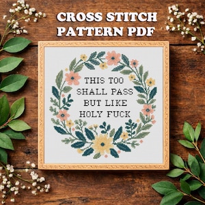 May include: A framed cross-stitch pattern PDF with a floral wreath design. The text within the wreath reads "THIS TOO SHALL PASS BUT LIKE HOLY FUCK". The frame is a light brown color, and the background is a wooden surface.