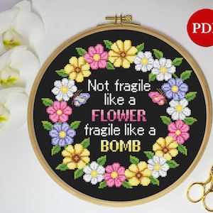 以下が含まれることがあります： 黒い背景に、カラフルな花輪と「Not fragile like a FLOWER fragile like a BOMB」というテキストが書かれたクロスステッチパターンです。