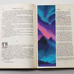 May include: An open book with a bookmark featuring a pixelated design of a mountain range under a colorful aurora borealis. The bookmark has shades of blue, purple, and pink. The book's pages are aged and filled with text.