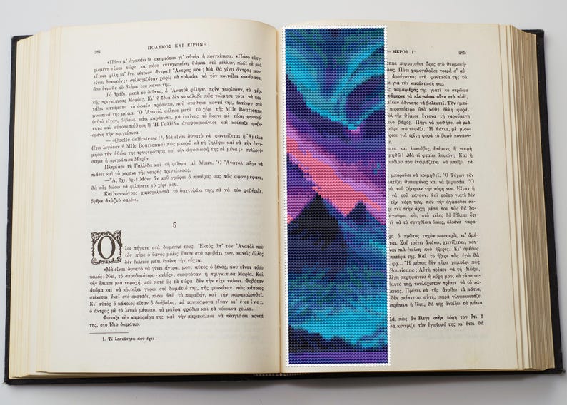 May include: An open book with a bookmark featuring a pixelated design of a mountain range under a colorful aurora borealis. The bookmark has shades of blue, purple, and pink. The book's pages are aged and filled with text.