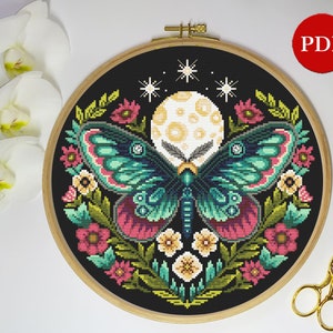 Puede incluir: Un patrón de punto de cruz con una mariposa verde y morada con una luna llena y estrellas en el fondo. La mariposa está rodeada de flores rosas, amarillas y verdes.