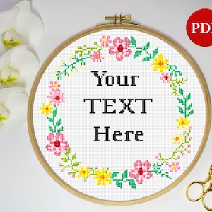 Può includere: Un cerchio da ricamo bianco con un design a corona di fiori in rosa, giallo e verde. Il testo "Your Text Here" è al centro del cerchio.