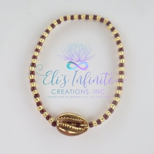 May include: A brown and gold beaded bracelet with a gold seashell charm. The bracelet is made with small, round beads and has a simple, elegant design.