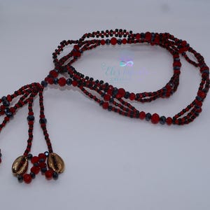 May include: A beaded necklace with red, black, and gray beads. The necklace has multiple strands and features gold-colored cowrie shell charms at the end. The design includes a logo with the text "Elis Talento Creations".