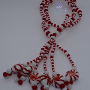 Op de afbeelding: Een ketting met rode en witte kralen. De ketting heeft meerdere strengen met snoepgoedvormige hangers en rood-witte swirl kralen. De tekst "Ele's Infinite Creations, INC" is zichtbaar.